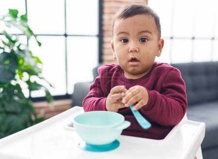 Photo for Adorable hispanic boy sitting on highchair holding spoon at home - Royalty Free Image