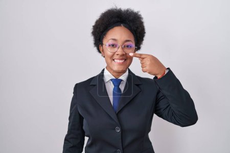 Téléchargez les photos : Belle femme africaine aux cheveux bouclés portant une veste d'affaires et des lunettes pointant du doigt vers le visage et le nez, souriante et joyeuse. concept de beauté - en image libre de droit
