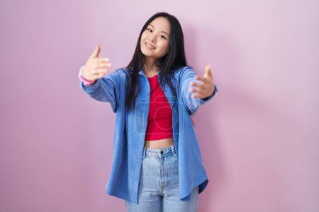 Téléchargez les photos : Young asian woman standing over pink background looking at the camera smiling with open arms for hug. cheerful expression embracing happiness. - en image libre de droit