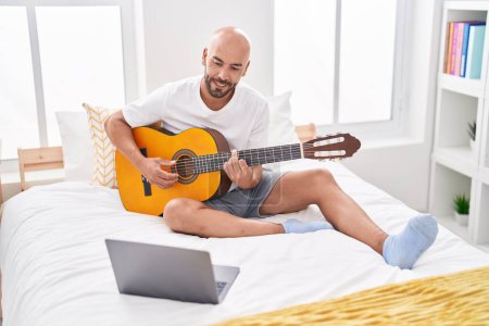 Photo for Young bald man having online classical guitar class sitting on bed at bedroom - Royalty Free Image