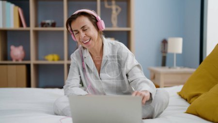Téléchargez les photos : Moyen Age femme hispanique écouter de la musique assis sur le lit dans la chambre - en image libre de droit