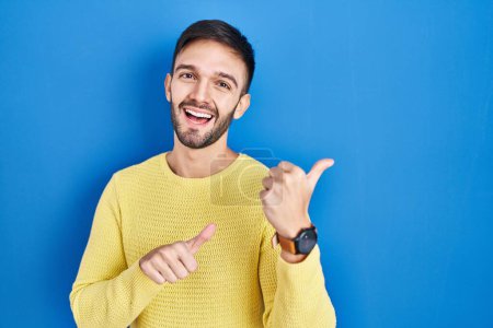 Foto de Hispanic man standing over blue background pointing to the back behind with hand and thumbs up, smiling confident - Imagen libre de derechos
