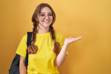 Foto de Joven mujer caucásica con mochila de estudiante sobre fondo amarillo sonriente alegre presentando y señalando con la palma de la mano mirando a la cámara. - Imagen libre de derechos