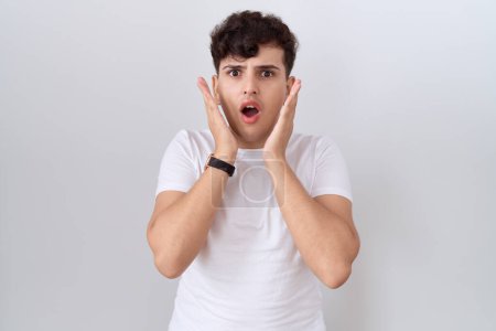 Téléchargez les photos : Jeune homme non binaire portant un t-shirt blanc décontracté effrayé et choqué, surprise et expression étonnante avec les mains sur le visage - en image libre de droit
