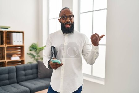 Foto de Hombre afroamericano trabajando en la oficina de terapia usando un teléfono inteligente apuntando el pulgar hacia el lado sonriendo feliz con la boca abierta - Imagen libre de derechos