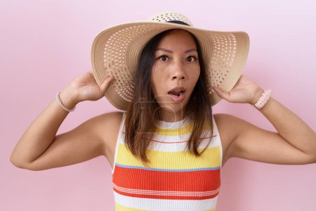 Photo for Middle age chinese woman wearing summer hat over pink background in shock face, looking skeptical and sarcastic, surprised with open mouth - Royalty Free Image