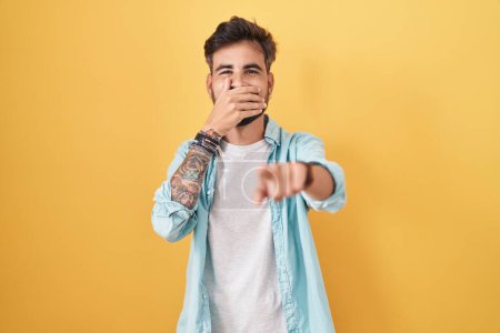 Foto de Young hispanic man with tattoos standing over yellow background laughing at you, pointing finger to the camera with hand over mouth, shame expression - Imagen libre de derechos