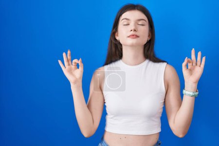 Téléchargez les photos : Jeune femme caucasienne debout sur fond bleu détendue et souriante avec les yeux fermés faisant geste de méditation avec les doigts. concept de yoga. - en image libre de droit
