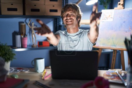 Téléchargez les photos : Homme d'âge moyen assis au studio d'art avec ordinateur portable la nuit en regardant la caméra souriant à bras ouverts pour étreindre. expression joyeuse embrassant le bonheur. - en image libre de droit