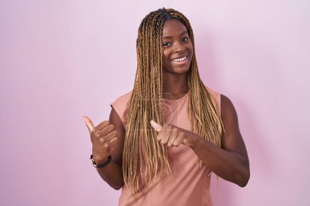 Téléchargez les photos : African american woman with braided hair standing over pink background pointing to the back behind with hand and thumbs up, smiling confident - en image libre de droit