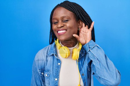 Foto de Mujer africana con trenzas de pie sobre fondo azul sonriendo con la mano sobre el oído escuchando y escuchando rumores o chismes. concepto de sordera. - Imagen libre de derechos