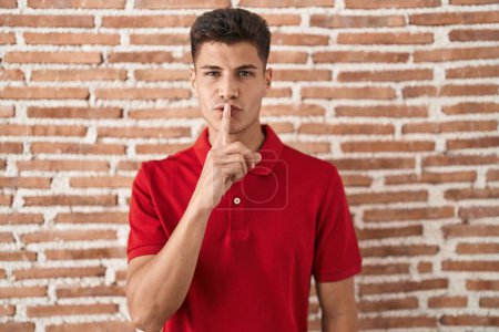 Téléchargez les photos : Jeune homme hispanique debout au-dessus d'un mur de briques demandant de se taire avec le doigt sur les lèvres. silence et concept secret. - en image libre de droit