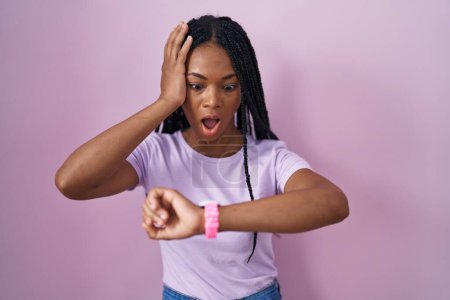 Téléchargez les photos : Femme afro-américaine avec des tresses debout sur fond rose regardant le temps de la montre inquiet, peur de se faire tard - en image libre de droit