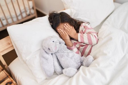 Téléchargez les photos : Adorable hispanique fille couchée sur le lit couvrant les yeux avec les mains à la chambre - en image libre de droit
