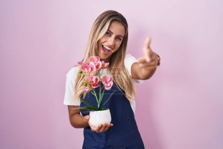Foto de Mujer rubia joven con delantal de jardinero sosteniendo la planta señalando los dedos a la cámara con la cara feliz y divertida. buena energía y vibraciones. - Imagen libre de derechos