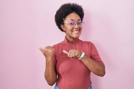 Foto de Hermosa mujer africana con el pelo rizado de pie sobre el fondo rosa apuntando a la espalda con la mano y los pulgares hacia arriba, sonriendo confiado - Imagen libre de derechos
