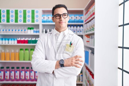 Téléchargez les photos : Young hispanic man pharmacist smiling confident standing with arms crossed gesture at pharmacy - en image libre de droit