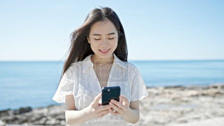 Téléchargez les photos : Jeune femme chinoise utilisant smartphone souriant au bord de la mer - en image libre de droit