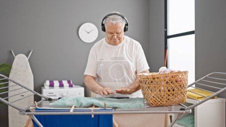 Photo for Middle age grey-haired man listening to music hanging clothes on clothesline at laundry room - Royalty Free Image