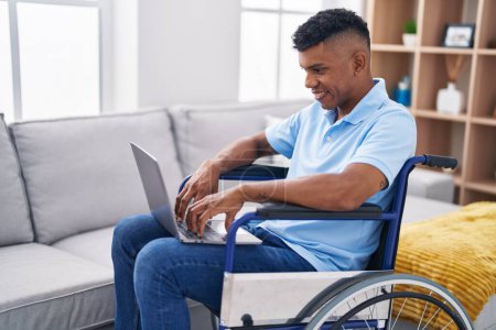 Foto de Hombre latino joven usando portátil sentado en silla de ruedas en casa - Imagen libre de derechos