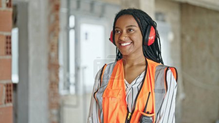 Téléchargez les photos : Femme afro-américaine constructeur souriant avec confiance casque avec bras croisés geste sur le chantier de construction - en image libre de droit