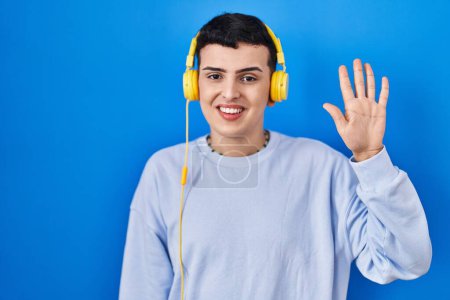 Foto de Persona no binaria escuchando música usando auriculares mostrando y apuntando hacia arriba con los dedos número cinco mientras sonríe confiado y feliz. - Imagen libre de derechos
