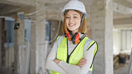 Foto de Joven mujer rubia arquitecta sonriendo confiada de pie con los brazos cruzados gesto en el sitio de construcción - Imagen libre de derechos