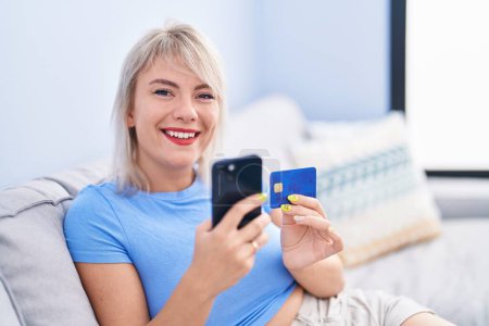 Téléchargez les photos : Jeune femme blonde utilisant smartphone et carte de crédit assis sur le canapé à la maison - en image libre de droit