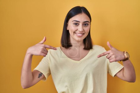 Foto de Chica hispana vistiendo camiseta casual sobre fondo amarillo mirando confiado con sonrisa en la cara, señalándose con los dedos orgullosos y felices. - Imagen libre de derechos