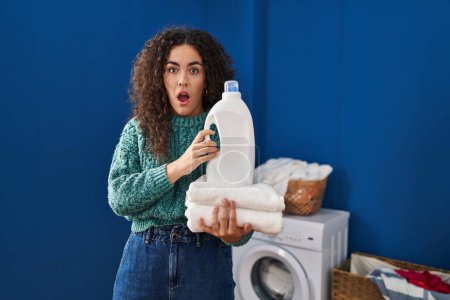 Photo for Young hispanic woman holding laundry and detergent bottle in shock face, looking skeptical and sarcastic, surprised with open mouth - Royalty Free Image
