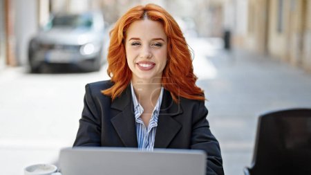 Foto de Joven pelirroja trabajadora de negocios usando laptop sonriendo en la terraza de la cafetería - Imagen libre de derechos