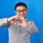 Young chinese man standing over blue background smiling in love doing heart symbol shape with hands. romantic concept.