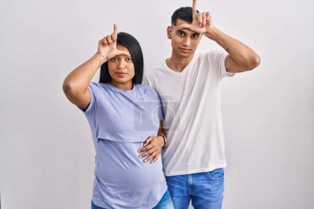 Photo for Young hispanic couple expecting a baby standing over background making fun of people with fingers on forehead doing loser gesture mocking and insulting. - Royalty Free Image