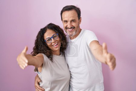 Téléchargez les photos : Couple hispanique moyen âge ensemble sur fond rose regardant la caméra souriant à bras ouverts pour étreindre. expression joyeuse embrassant le bonheur. - en image libre de droit