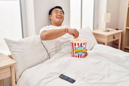 Téléchargez les photos : Jeune homme chinois regardant un film assis sur le lit dans la chambre - en image libre de droit