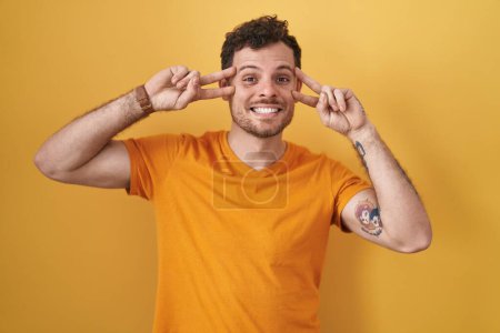 Foto de Joven hombre hispano de pie sobre fondo amarillo haciendo símbolo de paz con los dedos sobre la cara, sonriendo alegre mostrando la victoria - Imagen libre de derechos