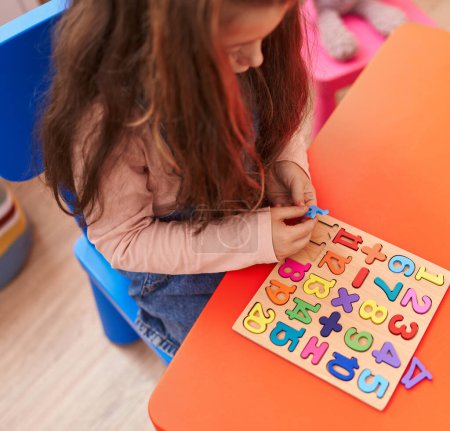 Foto de Adorable hispanic girl playing with maths puzzle game sitting on table at kindergarten - Imagen libre de derechos