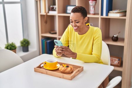 Photo for African american woman having breakfast using smartphone at home - Royalty Free Image