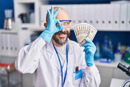 Foto de Joven hombre hispano trabajando en laboratorio científico sosteniendo dinero sonriendo feliz haciendo señal de bien con la mano en el ojo mirando a través de los dedos - Imagen libre de derechos