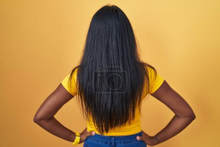 Téléchargez les photos : Jeune femme indienne debout sur fond jaune regardant vers l'arrière avec les bras sur le corps - en image libre de droit