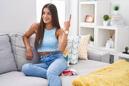 Téléchargez les photos : Jeune femme brune assise sur le canapé à la maison gaie avec un sourire sur le visage pointant avec la main et le doigt sur le côté avec une expression heureuse et naturelle - en image libre de droit
