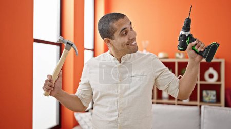 Foto de Joven hombre hispano sonriendo confiado sosteniendo taladro y martillo en casa - Imagen libre de derechos