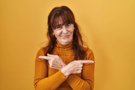 Foto de Mujer hispana de mediana edad de pie sobre fondo amarillo apuntando a ambos lados con los dedos, en diferente dirección en desacuerdo - Imagen libre de derechos
