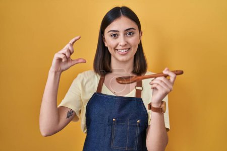 Photo for Hispanic girl eating healthy  wooden spoon smiling and confident gesturing with hand doing small size sign with fingers looking and the camera. measure concept. - Royalty Free Image