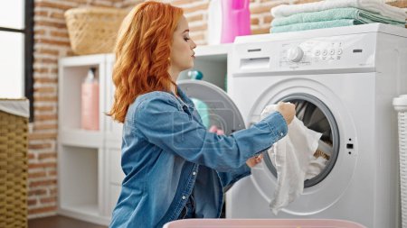 Foto de Joven pelirroja lavando ropa con expresión relajada en la lavandería - Imagen libre de derechos