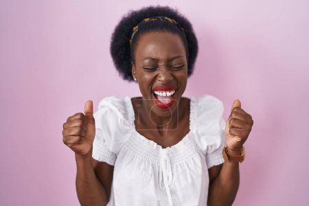 Téléchargez les photos : Femme africaine aux cheveux bouclés debout sur fond rose excité pour le succès avec les bras levés et les yeux fermés célébrant la victoire en souriant. concept gagnant. - en image libre de droit