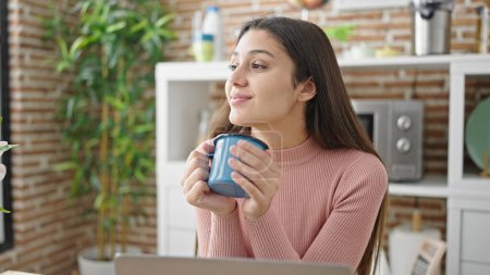 Photo for Young beautiful hispanic woman using laptop smelling cup of coffee at dinning room - Royalty Free Image