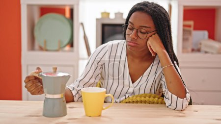 Téléchargez les photos : Femme afro-américaine versant du café sur une tasse assise sur une table fatiguée à la salle à manger - en image libre de droit