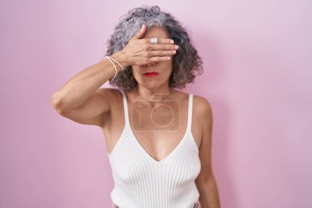 Foto de Middle age woman with grey hair standing over pink background covering eyes with hand, looking serious and sad. sightless, hiding and rejection concept - Imagen libre de derechos