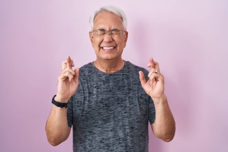 Téléchargez les photos : Homme du moyen âge avec les cheveux gris debout sur fond rose doigt gestuel croisé souriant avec espoir et les yeux fermés. chance et concept superstitieux. - en image libre de droit